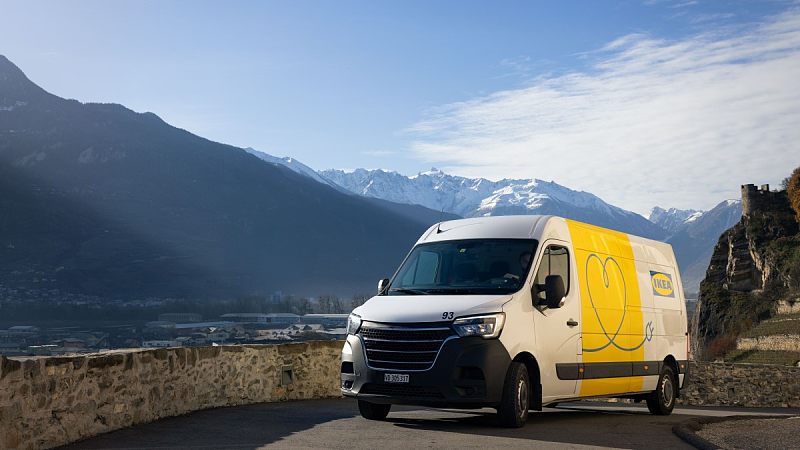 一辆宜家配送电动汽车行驶在瑞士的道路上，天空一片蔚蓝，背景中的山脉有一部分被白雪覆盖。