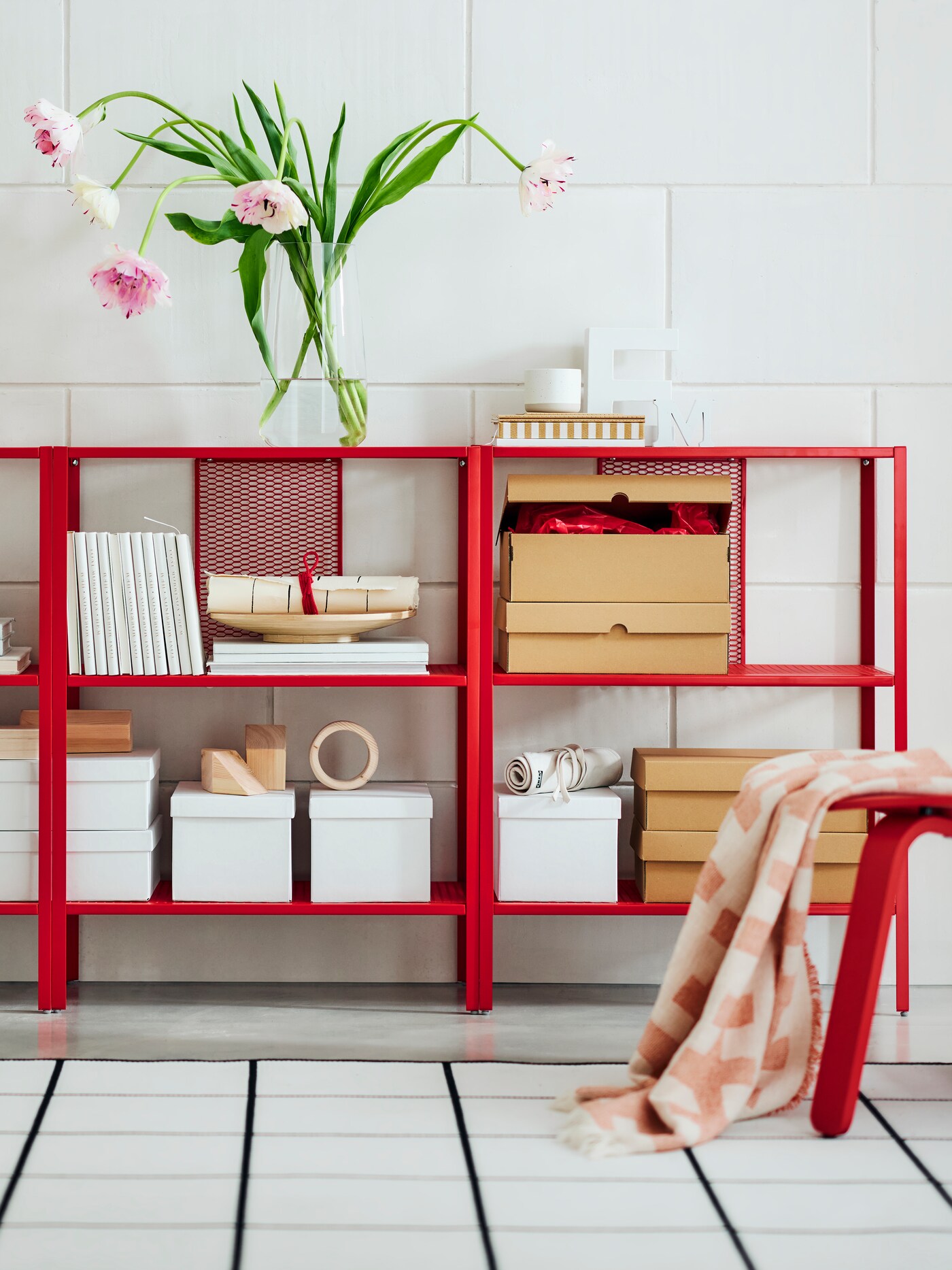 A bright red BAGGEBO shelving unit holding storage boxes leans against a white wall. Colourful flowers sit in a vase on top.