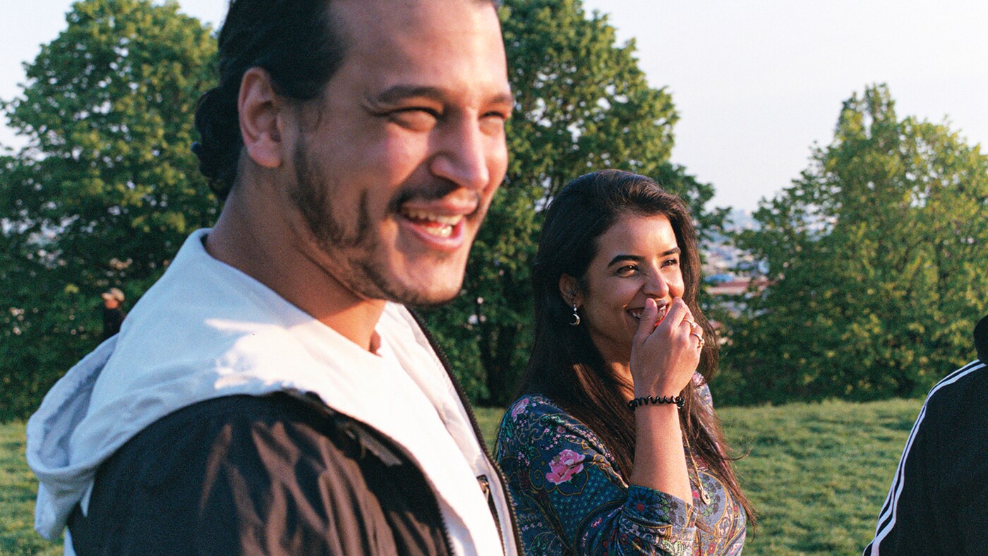 Image of a man and a woman laughing together, wearing casual clothes in front of trees in the middle of a field.