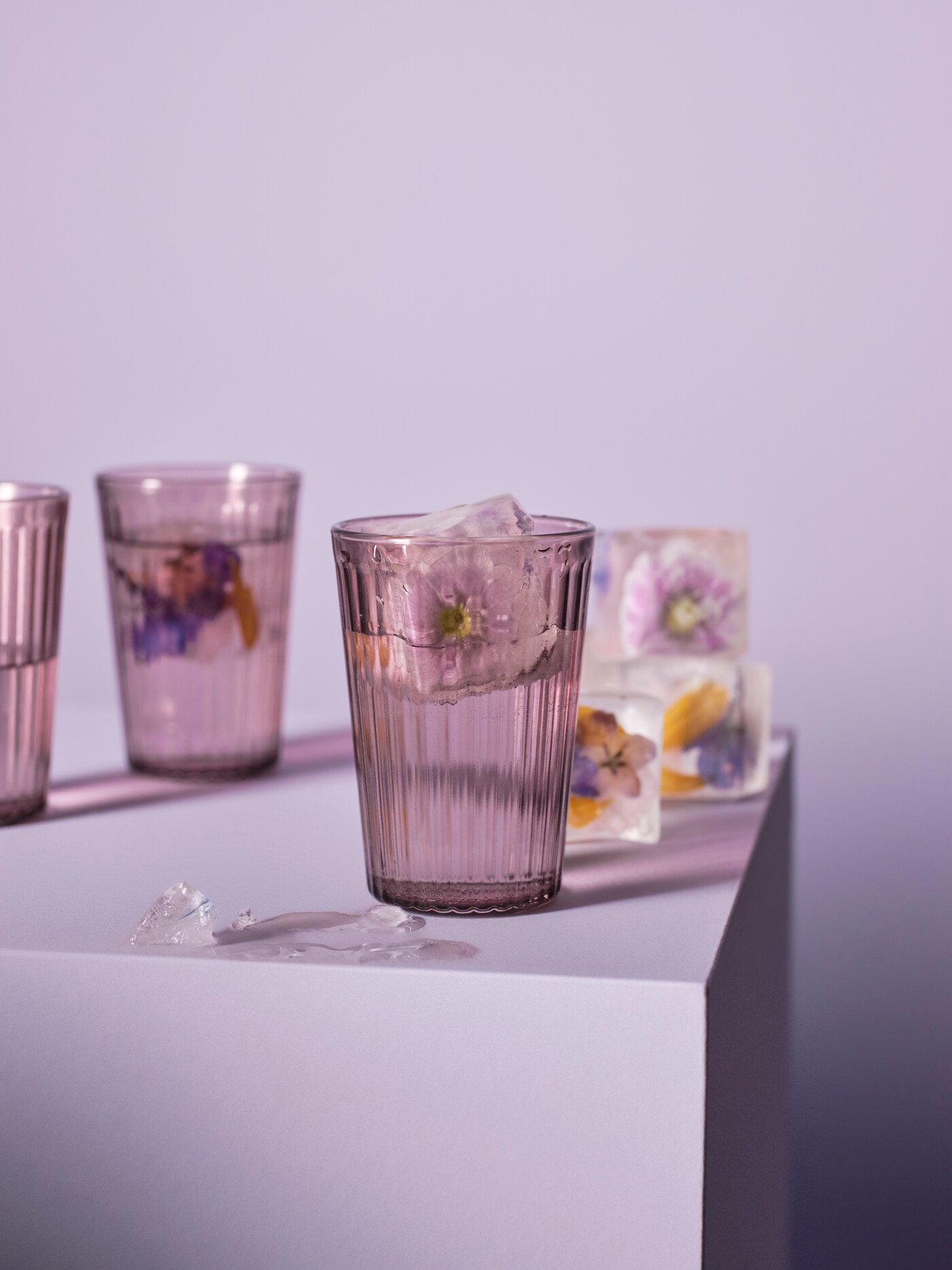 Three BROKROCKA grey-pink glasses sit on a lilac pedestal next to ice cubes with yellow and purple flowers frozen inside.